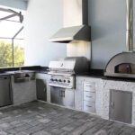 custom outdoor kitchen with stainless steel vent hood, grill, pizza oven, and cabinetry