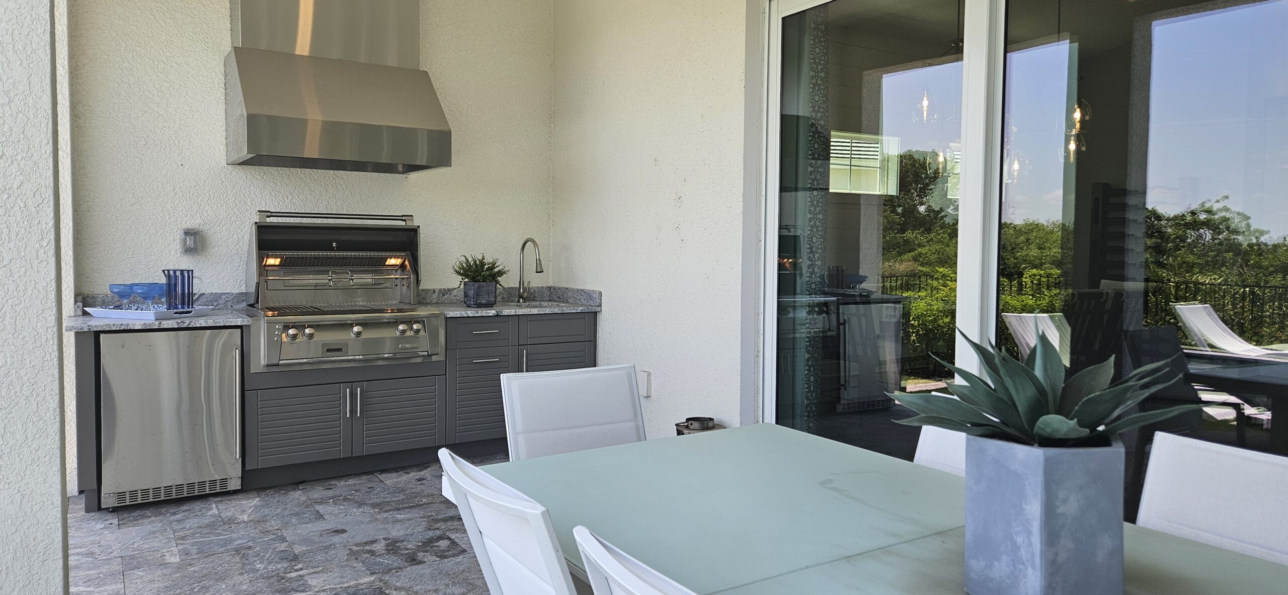 Outdoor patio with a stainless steel grill and a sink on a stone countertop. Elegant white dining set with a potted plant. Sunny, relaxed ambiance.